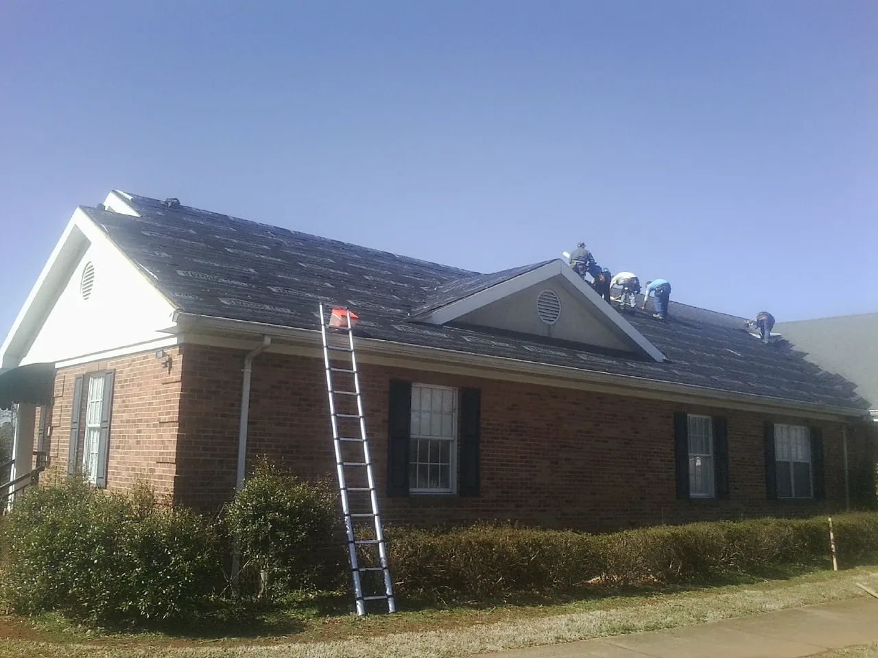 Crew tearing shingles off a brick ranch roof with a ladder set — Metal Roof Repair work by our crew in Glendale Springs