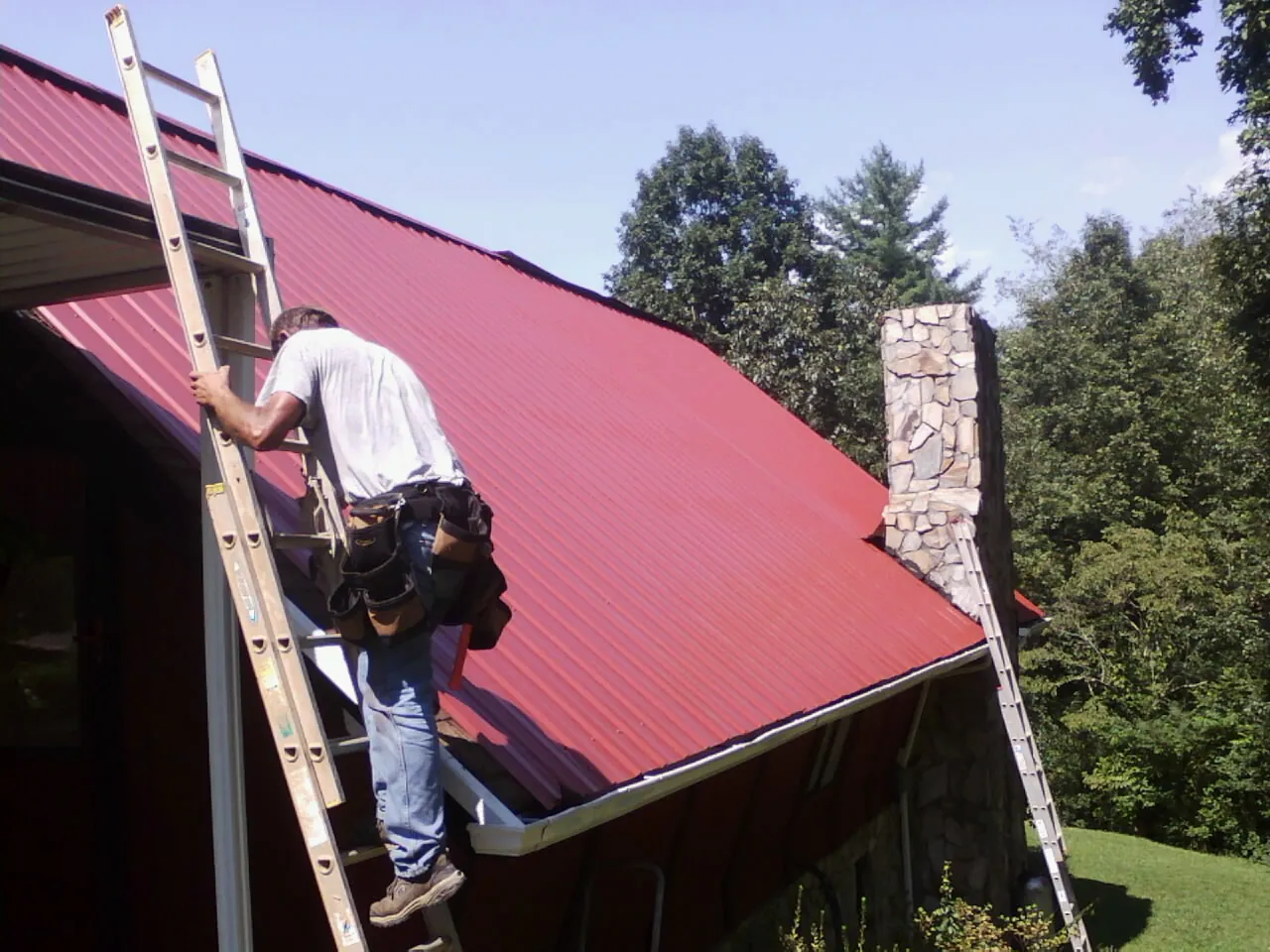 Roofer on a ladder beside a red metal roof and stone chimney — Soffit & Fascia Repair by Roofing Brothers in Glendale Springs